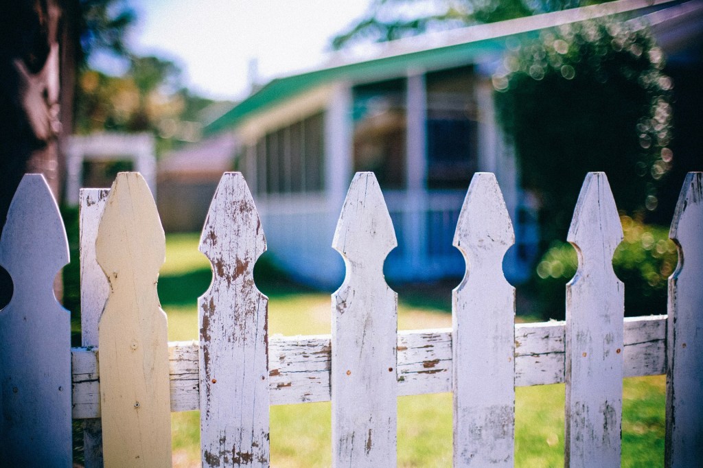 Faded paint on white picket fence in the foreground, grass and boka, blurred houses in the background in an urban neighborhood.