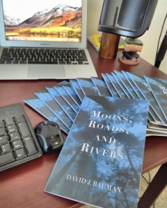 Poetry Chapbook Moons, Roads, and Rivers by David J. Bauman. Cover, midnight blue looking up through trees with moonlight. books, desk, mac, laptop, micraphone