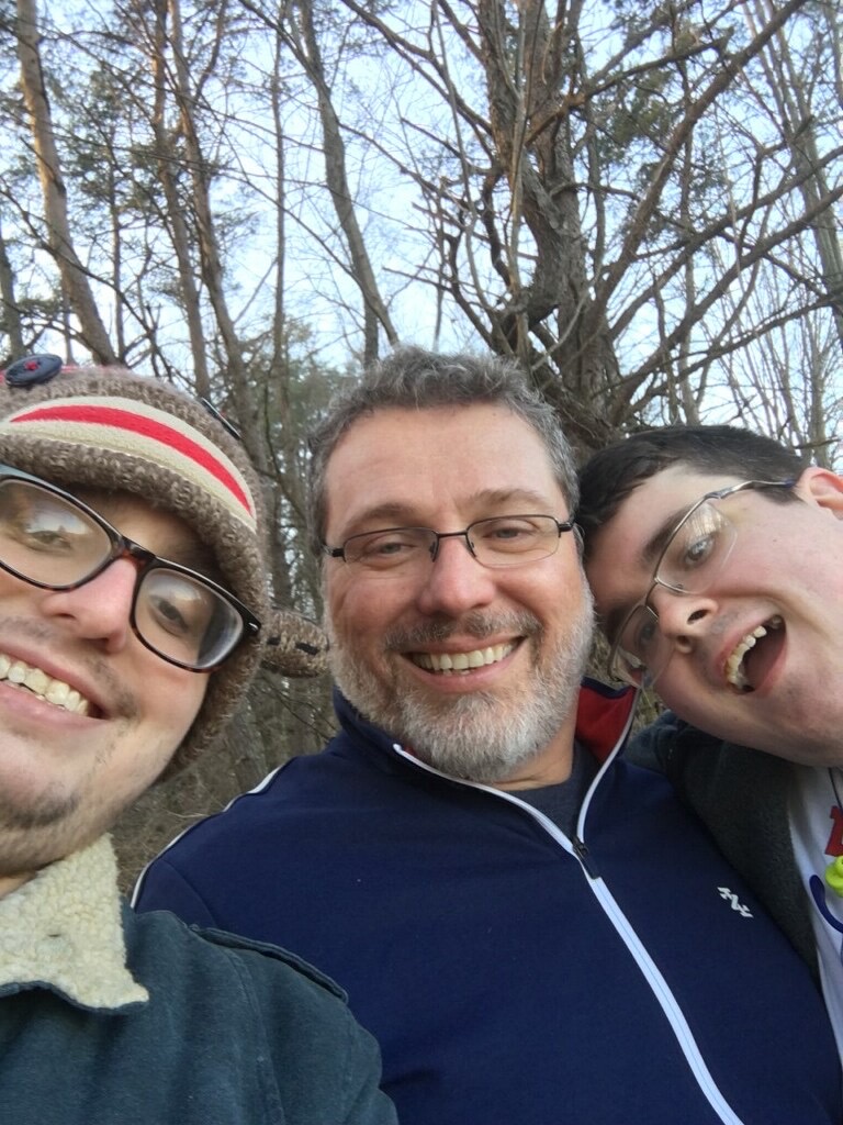 Three men smiling, winter hats and jackets, bare trees in the background